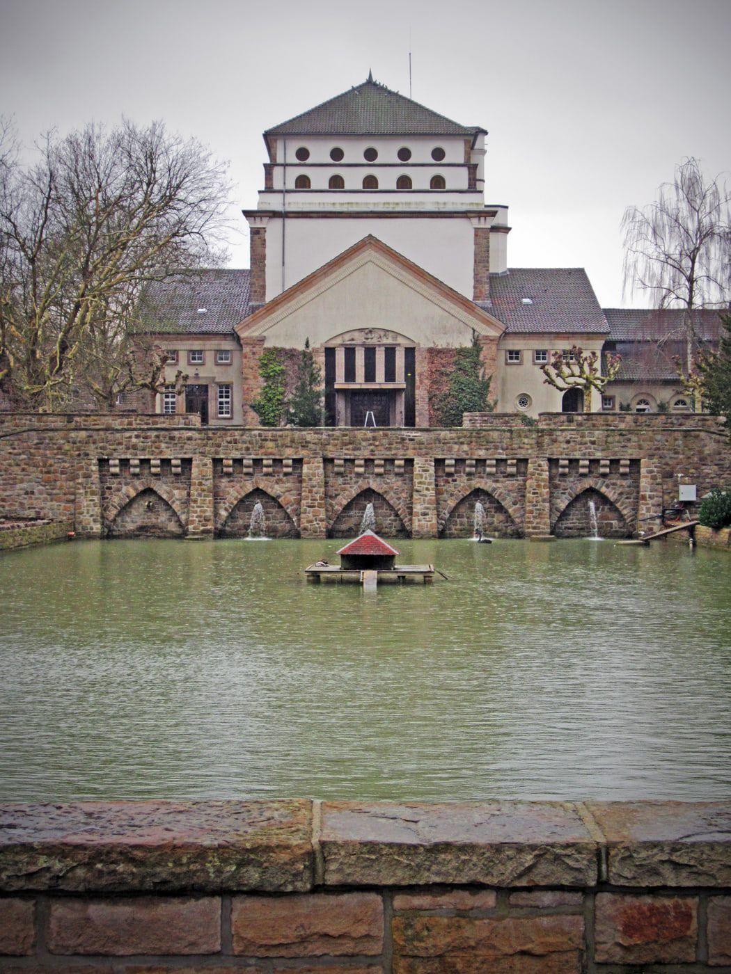Das expressionistische Hauptgebäude hinter einem großen Teich mit steinerner Bogenmauer, kleinen Fontänen und einem überdachten Podest im Wasser auf Hauptfriedhof Dortmund