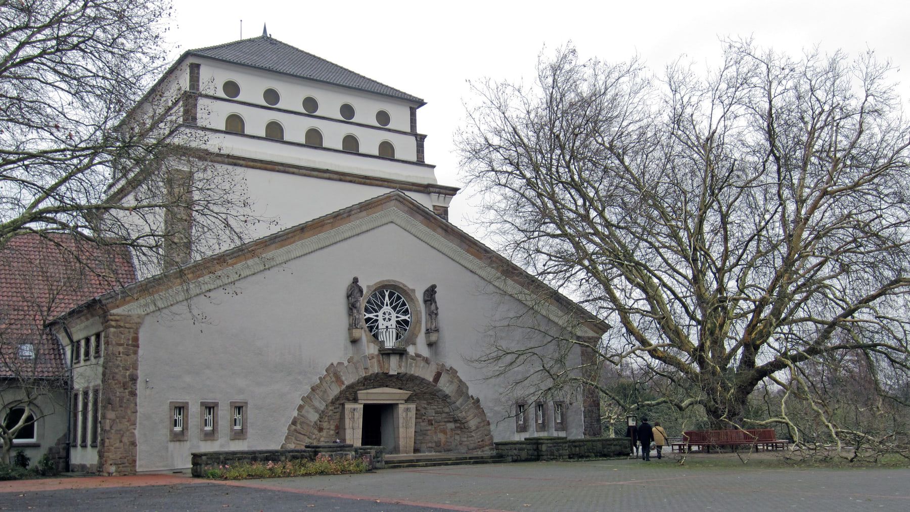 Schrägansicht des Hauptgebäudes mit Rundbogenportal und gestuftem Turm neben einem mächtigen laublosen Baum auf Hauptfriedhof Dortmund